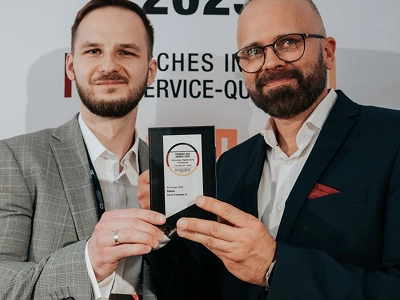Une large photographie montre la scène d'une cérémonie de remise de prix, prise depuis l'arrière d'un public assis. Sur le côté gauche de la scène, un homme en costume sombre et une femme en longue robe blanche se tiennent debout, une tablette à la main, face à la foule. Au centre de la scène, sur fond sombre, un grand écran affiche en haut un texte lumineux de couleur ambre qui dit « WIZJA I INNOVACJE ». En dessous, au centre de l'écran, se trouve un grand trophée en cristal en forme de diamant, suivi du texte « Maciej Góralski » et « Vasco Electronics » en dessous. Des lumières chaudes et diffuses éclairent la scène qui se détache sur l'auditorium sombre et bondé.