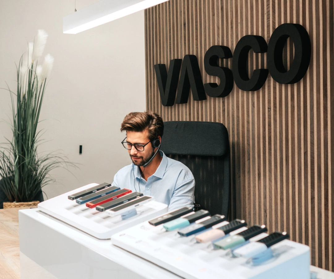 Un homme du service client est assis à un bureau affichant plusieurs appareils Vasco Translator de différentes couleurs, avec un grand panneau « VASCO » sur le mur en bois derrière lui.