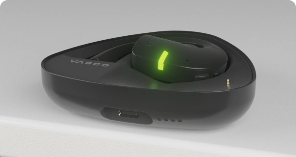 Un écouteur Vasco Translator E1 est placé dans son boîtier de recharge. Les voyants LED clignotants indiquent le niveau de batterie actuel du boîtier et de l'écouteur. 