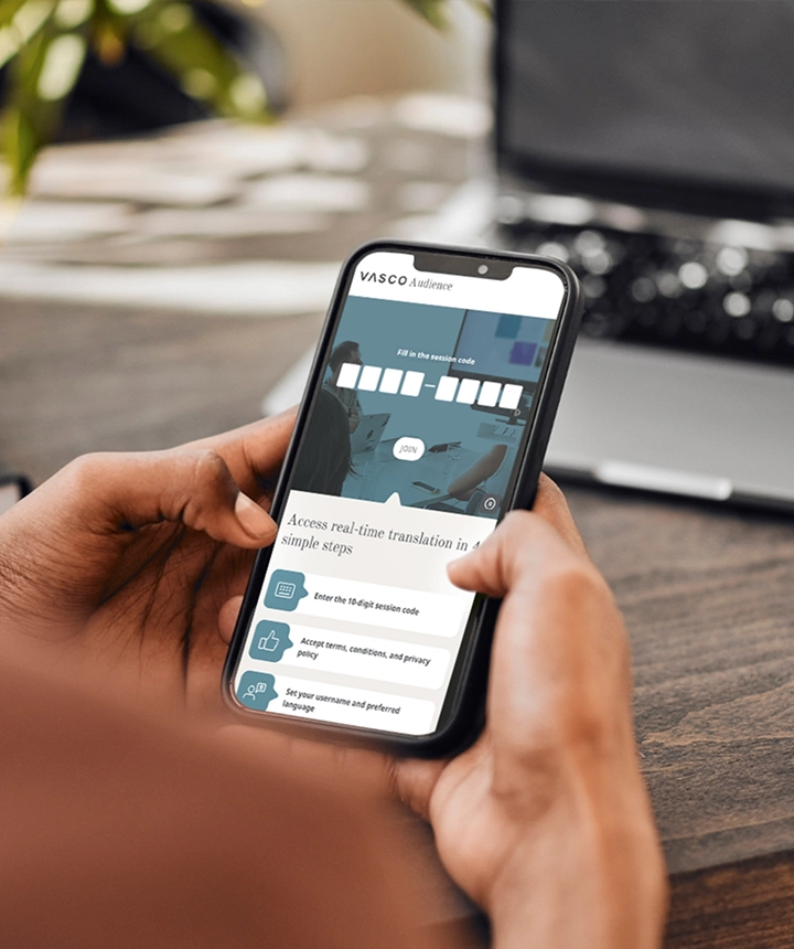 Vue par-dessus l'épaule d'une personne tenant un smartphone sur lequel s'affiche l'interface web de Vasco Audience. L'écran présente un champ de saisie intitulé « Saisissez le code de session » avec un bouton blanc « REJOINDRE », au-dessus d'une liste d'instructions indiquant « Accédez à la traduction en temps réel en 4 étapes simples ». À l'arrière-plan, on aperçoit un bureau en bois flou et un ordinateur portable. 