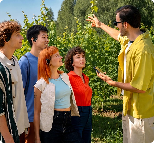 Un groupe de cinq personnes se trouve en plein air dans un vignoble baigné de soleil. À droite, un homme vêtu d'une chemise jaune et portant des lunettes de soleil tient un appareil de traduction portable Vasco Q1 et fait un geste en direction des vignes verdoyantes. À gauche, quatre personnes écoutent attentivement, chacune équipée d'un Vasco Translators E1 noir, illustrant ainsi la fonctionnalité de conversation de groupe en temps réel. 