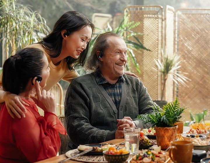 Une réunion familiale multigénérationnelle réconfortante autour d'une table débordant de mets. Un homme âgé souriant, vêtu d'un cardigan gris, et deux femmes discutent joyeusement dans une pièce baignée de soleil et remplie de plantes. Tous trois portent des écouteurs Vasco Translation E1 noirs. 