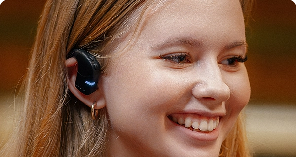 Gros plan sur le profil d'une jeune femme souriante portant un écouteur Vasco Translation E1 noir. L'image met en avant la conception hygiénique et ouverte de l'appareil, montrant qu'il se place confortablement à l'extérieur de l'oreille sans pénétrer dans le conduit auditif. 