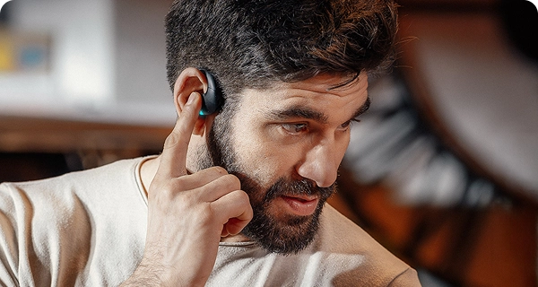 Gros plan sur un homme barbu qui tapote de l'index un écouteur de traduction Vasco noir placé sur son oreille, illustrant ainsi ses commandes tactiles intuitives. Une petite LED bleu vif est allumée sur l'appareil. 
