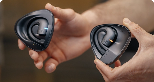 Gros plan sur des mains tenant deux étuis de recharge noirs distincts contenant des écouteurs Vasco avec fonction de traduction. L'image montre comment distinguer les deux modèles : l'un est équipé d'une LED bleue, tandis que l'autre dispose d'une LED orange, correspondant à des locuteurs différents. 