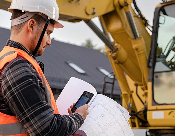 Un homme portant un casque et une tenue de travail prend une photo de plans de chantier à l'aide du Vasco Translator V4, avec une pelleteuse visible à l'arrière-plan.