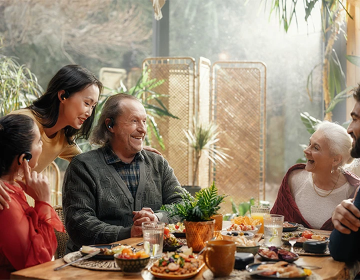 Une famille assise autour d'une table, équipée d'écouteurs Vasco Translator E1, qui rit et discute.