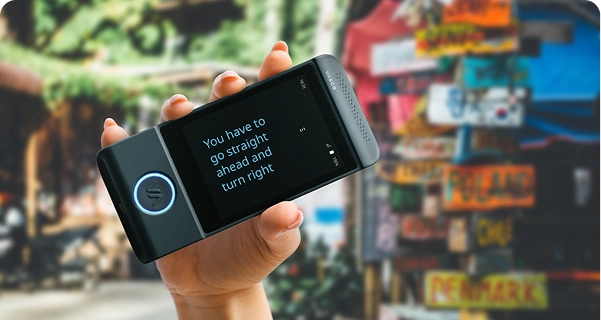 Gros plan sur une main tenant à l'horizontale un traducteur électronique Vasco noir. L'écran est en mode plein écran et affiche le texte traduit en grandes lettres bleues bien lisibles : « Vous devez continuer tout droit puis tourner à droite. » En arrière-plan, on aperçoit une scène extérieure floue et colorée, avec un poteau multicolore recouvert de panneaux indiquant la direction de différents pays, notamment la Pologne, le Chili et le Danemark. La lumière du soleil est vive et met en valeur le texte affiché sur l'appareil, facilitant ainsi la lecture. 