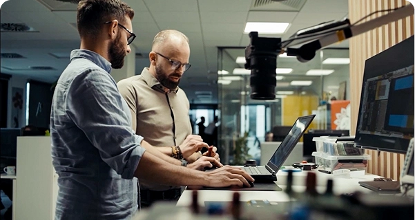 Deux hommes barbus et portant des lunettes travaillent ensemble devant un bureau debout, dans un bureau ou un laboratoire moderne. L'un d'eux, vêtu d'une chemise bleu clair, utilise un ordinateur portable, tandis que l'autre, vêtu d'une chemise beige, se tient à ses côtés et désigne d'un geste un petit composant électronique qu'il tient dans la main. Le bureau est équipé d'un grand écran, de divers outils électroniques et d'une caméra montée sur un bras articulé. En arrière-plan, on aperçoit un espace de travail lumineux et ouvert, délimité par des cloisons en verre. 
