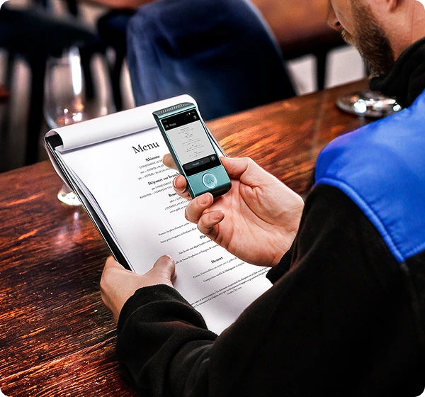 Gros plan en contre-plongée sur une personne assise à une table en bois dans un restaurant, utilisant un traducteur électronique portable pour lire un menu. La personne tient un menu papier blanc dans sa main gauche, tandis que de la main droite, elle tient au-dessus un petit traducteur Vasco de couleur bleu sarcelle. L'écran de l'appareil affiche une version numérique du menu en cours de traduction vers le polonais. La personne porte une veste noire avec un empiècement bleu vif sur l'épaule. L'arrière-plan est légèrement flou, laissant entrevoir l'intérieur chaleureux d'un restaurant. 