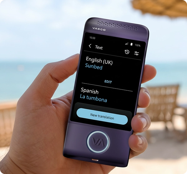 Gros plan sur une main tenant un traducteur électronique Vasco de couleur violette. L'écran affiche la traduction d'un texte de l'anglais (Royaume-Uni) vers l'espagnol, où le mot « Sunbed » est traduit par « La tumbona ». En arrière-plan, on aperçoit une plage ensoleillée avec une vue floue sur l'océan, du sable doré, plusieurs chaises longues et un parasol en paille. 