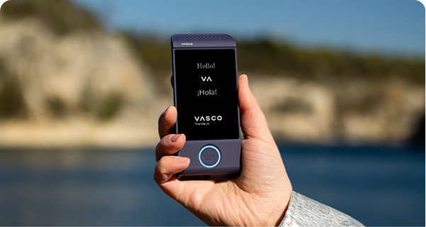 Gros plan sur une main tenant un traducteur électronique Vasco violet, sur fond de plein air lumineux. L'écran affiche les formules de salutation « Hello ! », « ¡Hola ! » et le logo « VASCO Translator ». L'arrière-plan présente une vue floue d'un paysage côtier ensoleillé, avec une mer bleue et des falaises rocheuses claires sous un ciel dégagé. La personne qui tient l'appareil porte une veste à manches texturées de couleur gris clair ou argenté. 