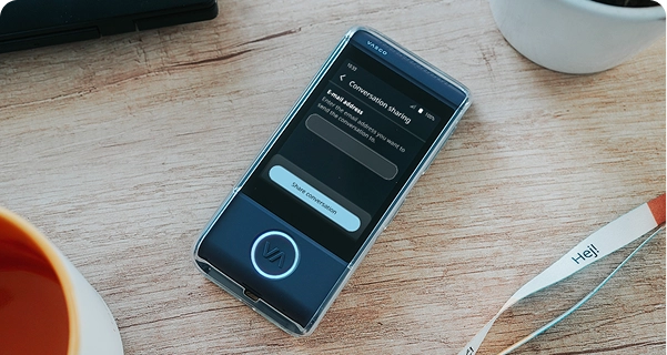 Un gros plan en contre-plongée d'un traducteur électronique Vasco bleu foncé, placé dans un étui de protection transparent, posé sur une table en bois de couleur claire. L'écran de l'appareil est allumé et affiche un menu « Partage de conversation » comportant un champ pour saisir une adresse e-mail et un bouton « Partager la conversation ». Autour de l'appareil, sur la table, on aperçoit le bord d'une tasse à café orange, un pot de fleur blanc et un cordon blanc sur lequel est imprimé le mot « Hej ! ». La scène est très bien éclairée, ce qui crée une esthétique épurée et moderne. 