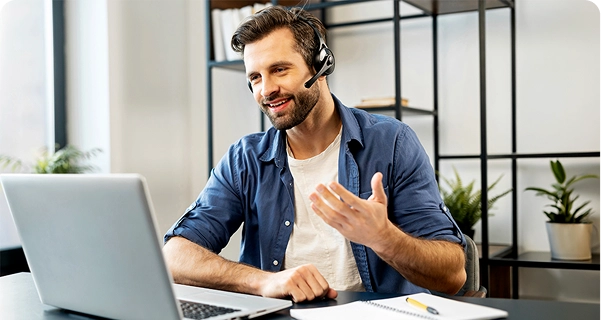 Un homme à l'air sympathique, barbu et aux cheveux foncés, est assis à un bureau. Il porte un casque professionnel muni d'un micro. Il sourit et gesticule d'une main tout en regardant l'écran d'un ordinateur portable ; il semble être en plein appel vidéo ou en train d'apporter une assistance technique. Il porte une chemise bleue boutonnée par-dessus un t-shirt blanc. Sur le bureau devant lui se trouvent un ordinateur portable, un cahier et un stylo jaune. L'arrière-plan représente un espace de bureau lumineux et moderne, avec une étagère minimaliste en métal noir et des plantes en pot vertes. 