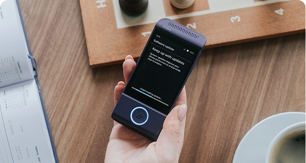 Une vue plongeante sur la main d'une personne tenant un traducteur électronique Vasco violet au-dessus d'une table en bois. L'écran de l'appareil affiche un message intitulé « Mise à jour logicielle » avec la mention « Restez à jour », ainsi qu'une barre de progression indiquant « Téléchargement de la mise à jour... ». Sur la table, autour de l'appareil, se trouvent un échiquier en bois avec des pièces d'échecs, un calendrier ouvert et une tasse de café blanche. L'éclairage est doux et naturel, créant une atmosphère détendue et quotidienne. 