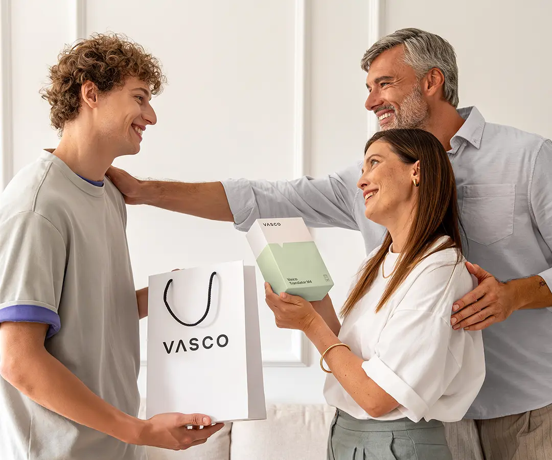 Une scène réconfortante où l'on voit trois personnes debout, souriantes, côte à côte. À gauche, un jeune homme aux cheveux bouclés tient un sac de courses blanc « VASCO ». Au centre, une femme tient une boîte blanche et vert clair portant l'inscription « Vasco Translator M4 » et regarde le jeune homme avec un air heureux. Derrière elle, un homme aux cheveux grisonnants sourit chaleureusement, une main posée sur l'épaule de la femme et l'autre sur celle du jeune homme. Ils se trouvent dans une pièce lumineuse aux murs de couleur neutre, avec un canapé en arrière-plan. 
