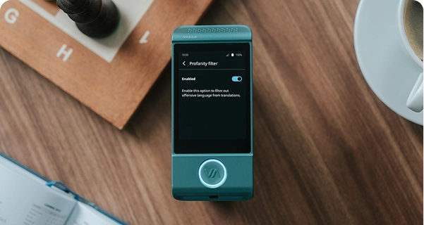 Une vue plongeante d'un traducteur électronique Vasco de couleur bleu sarcelle posé sur une table en bois. L'écran de l'appareil est allumé et affiche le menu des paramètres du « filtre anti-grossièreté », dont le bouton est réglé sur « Activé ». Sous le bouton, une description indique : « Activez cette option pour filtrer les propos grossiers dans les traductions. » L'appareil est entouré d'objets du quotidien, notamment un échiquier en bois avec une pièce de couleur sombre, un coin d'un cahier ouvert et une tasse de café blanche, le tout baigné d'une lumière douce et naturelle. 