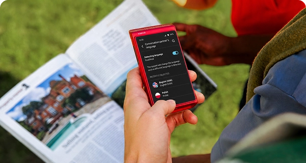 Une personne utilise un Vasco Translator Q1 rouge pour prendre une photo d'un journal. L'écran affiche le menu de sélection de la langue permettant de traduire le texte détecté automatiquement dans le journal.