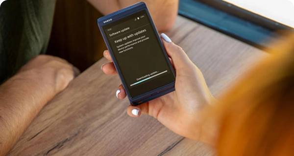 Vue par-dessus l'épaule d'une femme tenant un Vasco Translator Q1 bleu au-dessus d'une table en bois à l'intérieur d'un train. L'écran de l'appareil affiche une notification « Mise à jour logicielle » accompagnée d'une barre de progression indiquant « Téléchargement de la mise à jour... ».